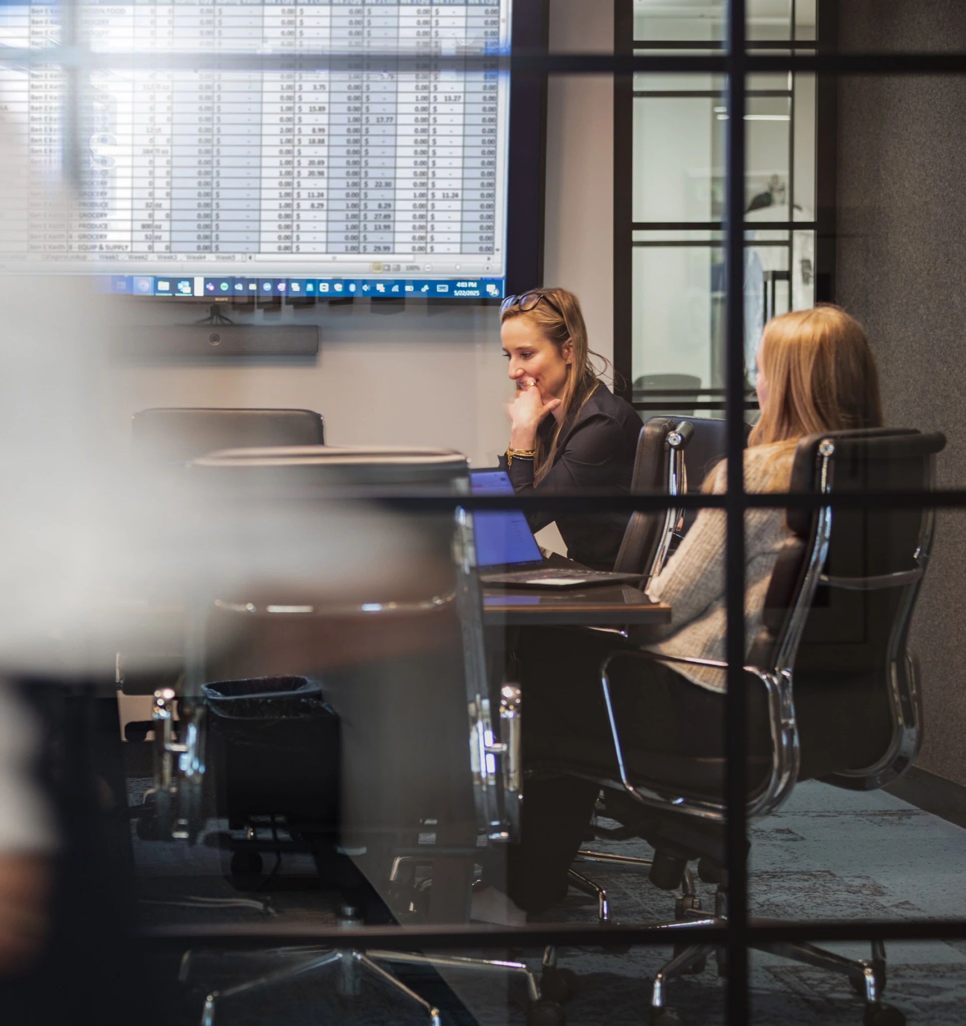 Two women collaborating in corporate boardroom while analyzing numbers projected on a digital monitor