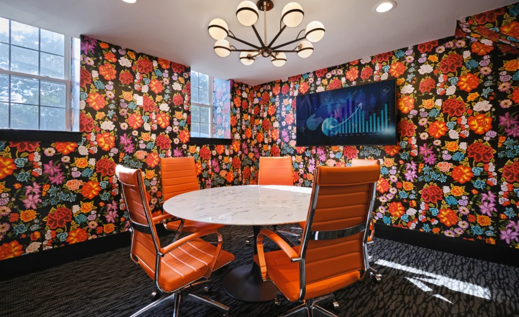 Vibrant meeting room with floral wallpaper, round table, orange chairs, and mounted TV screen