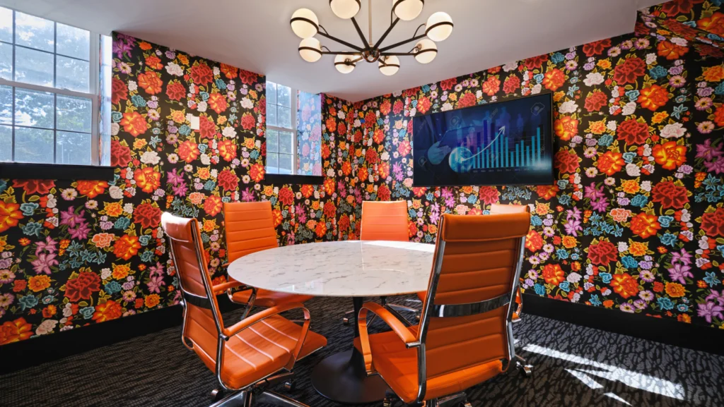 Vibrant meeting room with floral wallpaper, round table, orange chairs, and mounted TV screen