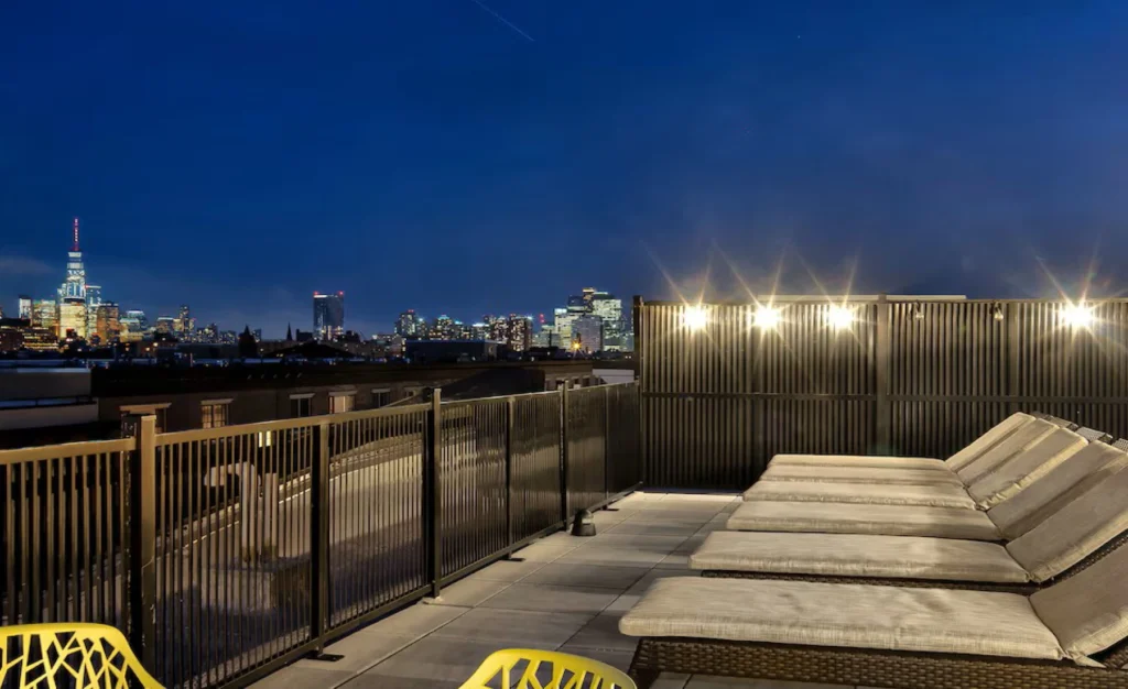 Rooftop lounge with cushioned sunbeds overlooking city skyline under evening lights at Halstead Hoboken
